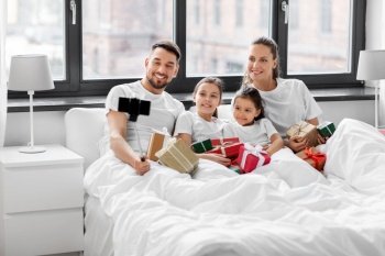 family, winter holidays and people concept - happy mother, father and two daughters with christmas gifts taking selfie on smartphone in bed at home. family with christmas gifts taking selfie in bed