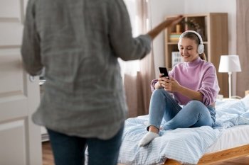 misbehavior, conflict and family concept - angry mother entering room and smiling daughter in headphones with smartphone sitting on bed at home. angry mother and daughter with smartphone at home