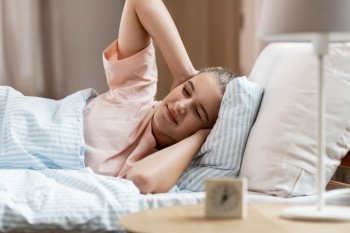 people, bedtime and rest concept - sleepy girl with alarm clock awaking in bed at home. sleepy girl with alarm clock awaking in bed