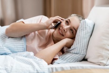 people, bedtime and rest concept - happy smiling girl calling on smartphone lying in bed at home. happy girl calling on smartphone in bed at home