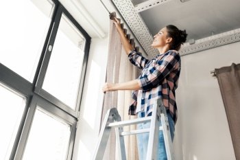 home improvement, decoration and renovation concept - happy smiling woman on ladder hanging curtains. woman on ladder hanging curtains at home
