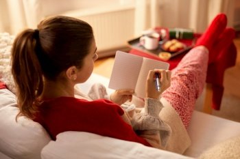 christmas, winter holidays and leisure concept - close up of young woman with pencil writing to notebook and resting her feet on table at cozy home. woman writing to notebook at home on christmas