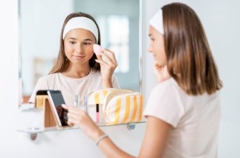 beauty, make up and cosmetics concept - teenage girl with sponge applying foundation to face and using smartphone in front of mirror at home bathroom. teenage girl with smartphone using make up sponge