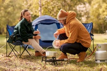 camping, tourism and travel concept - happy couple drinking beer and cooking food in pot on tourist gas burner at tent camp. couple drinking beer and cooking food at tent camp