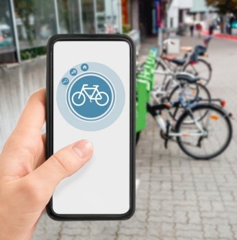 technology, transport and sustainability concept - close up of hand holding smartphone with mobile app and bicycle icon on screen over electric bike parking and charging station in city on background. hand with phone at electric bike charging station