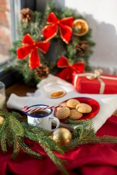 christmas and winter holidays concept - camp cup of coffee with candy cane, oatmeal cookies on red ceramic plate, gift and decorations on window sill at home. cup of coffee, cookies and christmas decor at home