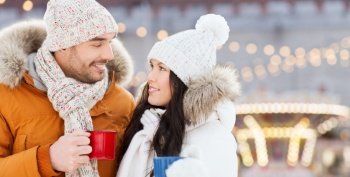 people, winter holidays and leisure concept - happy couple holding hot tea cups over christmas lights in amusement park on background. happy couple with tea cups over christmas lights