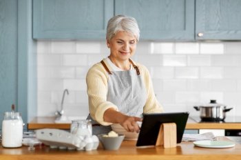 culinary, baking and technology concept - happy smiling woman looking at tablet pc computer cooking food on kitchen at home. woman with tablet computer cooking food on kitchen
