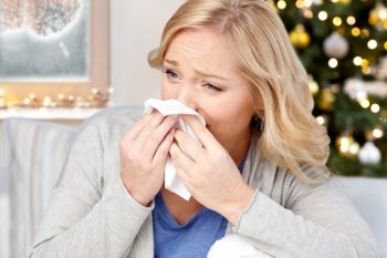 health, winter holidays and people concept - ill middle aged woman blowing nose to paper tissue at home over christmas tree lights on background. woman blowing nose to paper tissue on christmas