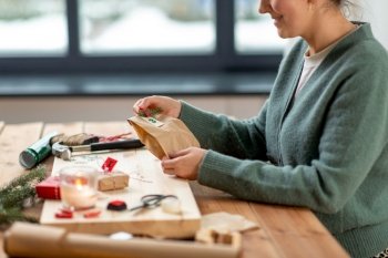 christmas, winter holidays and hobby concept - close up of woman with clothespin, craft paper bag and tag packing gift and making advent calendar at home. woman making christmas advent calendar at home