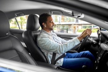 transport, people and technology concept - smiling indian man or driver with wireless earphones or hands free device driving car. man or driver with wireless earphones driving car