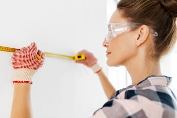 repair, construction and building concept - woman in goggles and protective gloves with ruler and pencil measuring wall at home. woman with ruler and pencil measuring wall at home