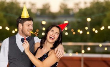birthday, celebration and holidays concept - happy couple with party blowers and caps having fun at rooftop party over lights on background. happy couple with party blowers having fun