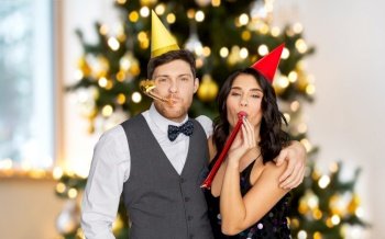 celebration and winter holidays concept - happy couple with party blowers and caps having fun over christmas tree lights background. happy couple with party blowers on christmas