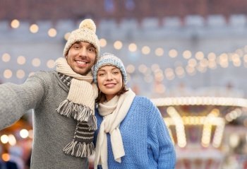 people and winter holidays concept - happy couple in knitted hats and scarves taking selfie over christmas market and lights background. happy couple in winter clothes taking selfie