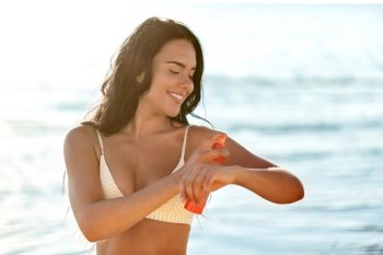people, summer and swimwear concept - happy smiling young woman in bikini swimsuit using sunscreen spray on beach. smiling woman in bikini with sunscreen on beach