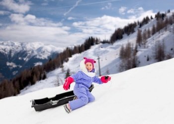 childhood, leisure and winter season concept - happy little girl with sled on snow hill over snow-covered mountains or alps on background. little girl with sled on snow hill in winter