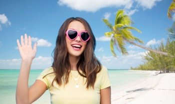 summer holidays, valentine’s day and travel concept - smiling young woman or teenage girl in heart-shaped sunglasses waving hand over tropical beach background in french polynesia. happy woman in heart-shaped sunglasses on beach