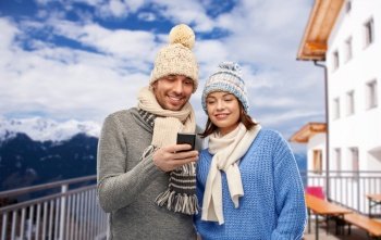 travel, tourism and winter holidays concept - happy couple in knitted hats and scarves with smartphone over mountains and ski resort background. happy couple with smartphone in winter