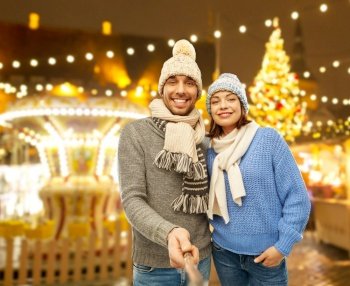 people, technology and winter holidays concept - happy couple in knitted hats and scarves taking picture by selfie stick over night christmas market and lights background. happy couple in winter clothes taking selfie