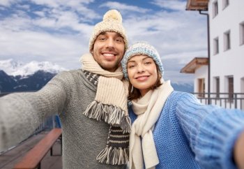 travel, tourism and winter holidays concept - happy couple in knitted hats and scarves taking selfie over mountains and ski resort background. happy couple taking selfie in winter