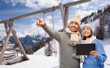 travel, tourism and winter holidays concept - happy couple in knitted hats and scarves with tablet computer over mountains and ski resort background. couple with tablet computer in winter