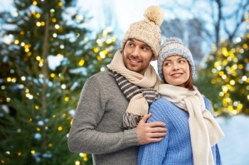 people and winter holidays concept - happy couple in knitted hats and scarves outdoors over christmas tree lights background. happy couple in winter clothes