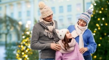 family, people and winter holidays concept - happy mother, father and little daughter in knitted hats and scarves over christmas tree lights on city street background. happy family in winter clothes on grey background