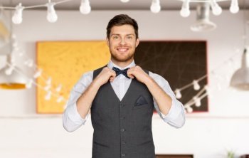 fashion, style and holidays concept - happy man in festive suit dressing for party and adjusting bowtie over restaurant background. happy man in festive suit dressing for party