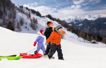 childhood, sledging and season concept - group of happy little kids walking, with sleds in winter over alps mountains on background. happy little kids walking with sleds in winter