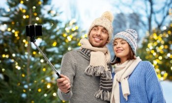 people, technology and winter holidays concept - happy couple in knitted hats and scarves taking picture by smartphone on selfie stick outdoors over christmas tree lights background. happy couple in winter clothes taking selfie