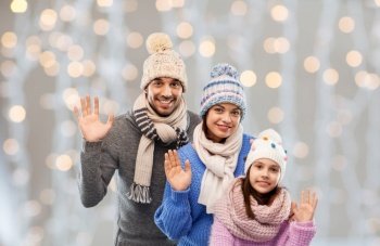 family, people and winter holidays concept - happy mother, father and little daughter in knitted hats and scarves waving hands over christmas lights background. happy family in winter clothes waving hands
