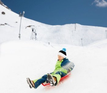 childhood, sledging and season concept - happy little boy sliding on sleds down snow hill in winter over alps mountains on background. happy boy sliding on sleds down hill in winter
