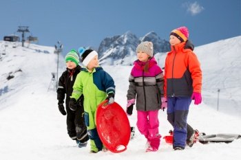 childhood, sledging and season concept - group of happy little children with sleds in winter over alps mountains on background. happy little children with sleds in winter