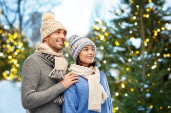 people and winter holidays concept - happy couple in knitted hats and scarves outdoors over christmas tree lights outdoors background. happy couple in winter clothes