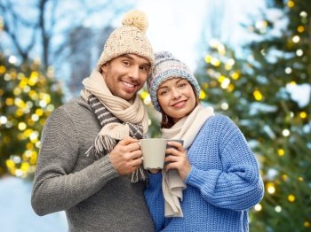 people and winter holidays concept - happy romantic couple in knitted hats and scarves with mugs drinking coffee outdoors over christmas tree lights background. happy couple in winter clothes with mugs