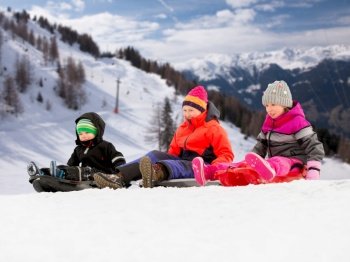 childhood, sledging and season concept - group of happy little kids sliding on sleds in winter over alps mountains on background. happy little kids sliding on sleds in winter