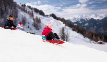 childhood, sledging and season concept - group of happy little kids sliding on sleds down snow hill in winter over alps mountains on background. kids sliding on sleds down snow hill in winter