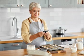 cooking, food and culinary concept - happy smiling senior woman with smartphone taking picture of cupcakes in baking mold on kitchen at home. woman with smartphone and cupcakes on kitchen