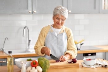 healthy eating, food cooking and culinary concept - happy smiling senior woman with knife chopping red onion on kitchen at home. happy woman chopping red onion on kitchen