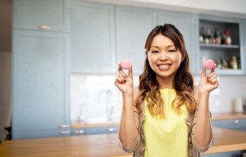 food, cooking and baking concept - happy asian young woman with macarons at home over kitchen background. happy woman with macarons on kitchen