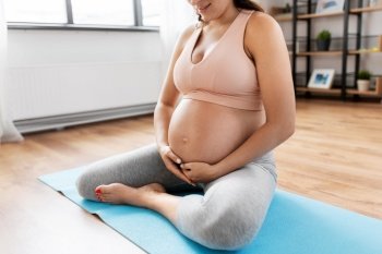 pregnancy, sport and fitness concept - close up of happy pregnant woman sitting on yoga mat at home and touching her belly. close up of pregnant woman doing yoga at home