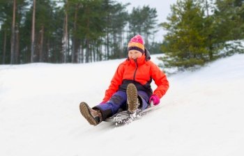 childhood, sledging and season concept - happy little girl sliding down on snow saucer sled outdoors in winter over snowy park or forest background. girl sliding down on snow saucer sled in winter