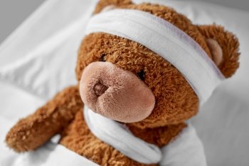 medicine, healthcare and childhood concept - ill teddy bear toy with bandaged paw and head lying in bed. ill teddy bear with bandaged paw and head in bed
