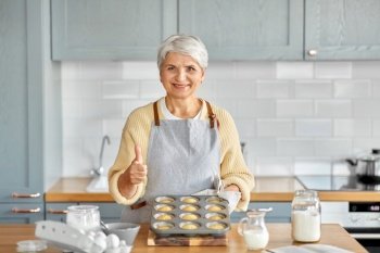 cooking, food and culinary concept - happy smiling senior woman with cupcakes in baking mold showing thumbs up on kitchen at home. woman with cupcakes in baking mold on kitchen