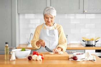healthy eating, food cooking and culinary concept - happy smiling senior woman with knife chopping champignons on kitchen at home. happy woman chopping champignons on kitchen