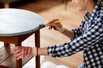 furniture renovation, diy and home improvement concept - close up of woman in gloves with paint brush painting old wooden table in grey color. woman painting old wooden table in grey color