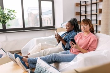 moving, people and real estate concept - happy smiling women with smartphones and boxes at new home. women with smartphones moving into new home