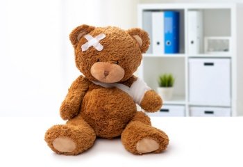 medicine, healthcare and childhood concept - teddy bear toy with bandaged paw and patch on head over medical office at hospital background. teddy bear toy with bandaged paw and patch on head