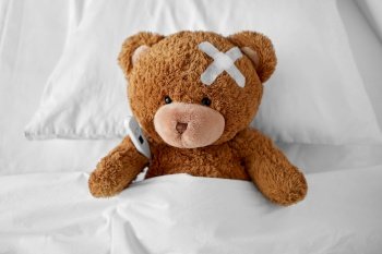 medicine, healthcare and childhood concept - ill teddy bear toy with medical patch on head and thermometer lying in bed. ill teddy bear with patch and thermometer in bed